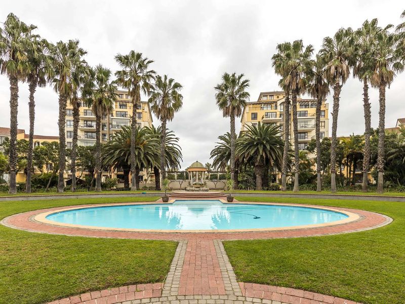 A well-maintained communal pool area featuring a circular swimming pool, brick pathways, and mature palm trees set within a landscaped garden.
