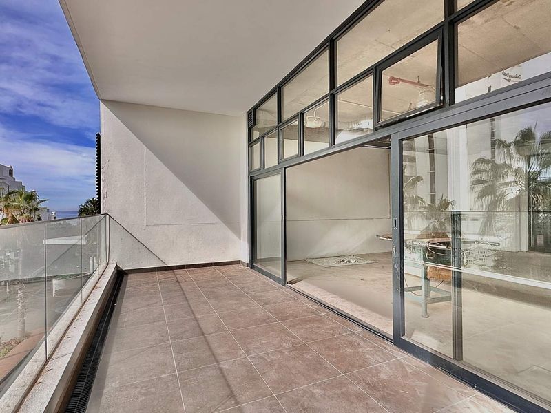 A spacious covered balcony featuring tiled flooring and a modern glass balustrade, providing seamless indoor-outdoor flow through large sliding glass doors.