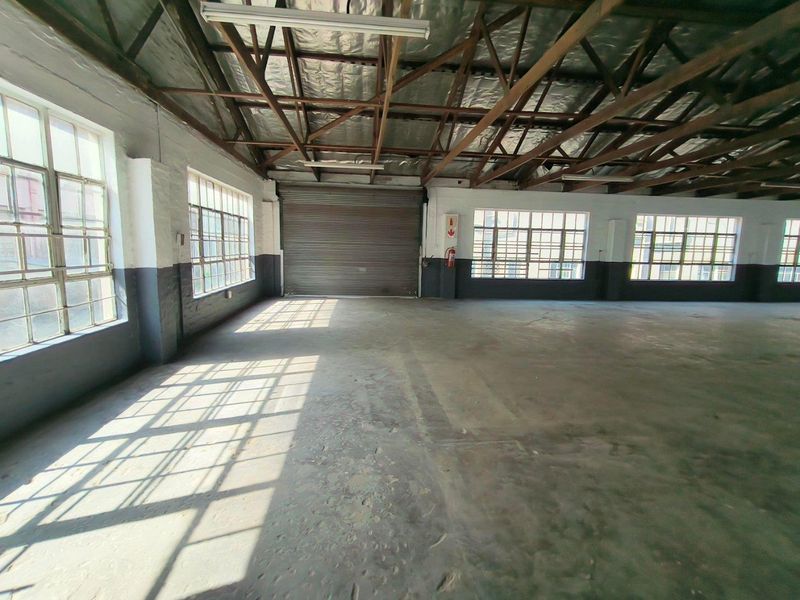 Spacious industrial warehouse unit featuring exposed steel roof trusses, large steel-framed windows for natural light, and a functional roller shutter door.