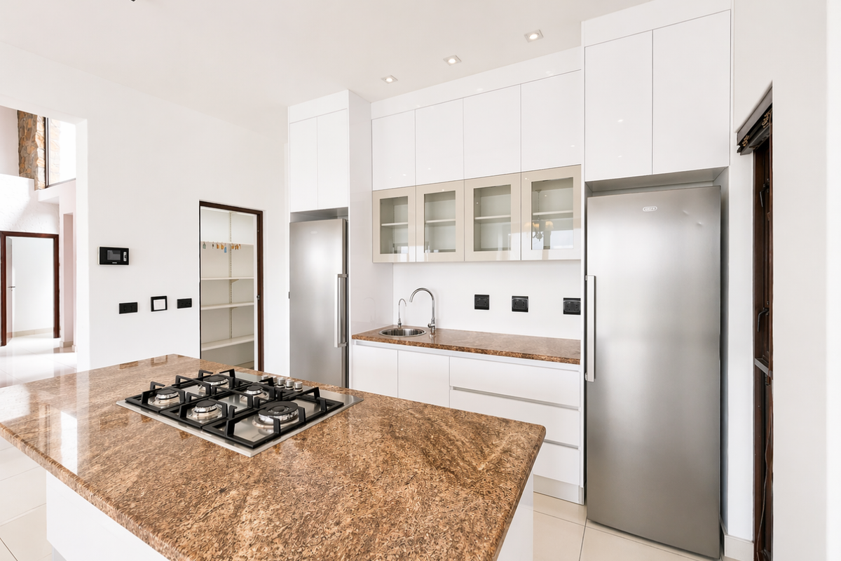 Modern kitchen featuring a large granite-topped island with a built-in gas hob, sleek white cabinetry, stainless steel appliances, and an adjacent walk-in pantry.