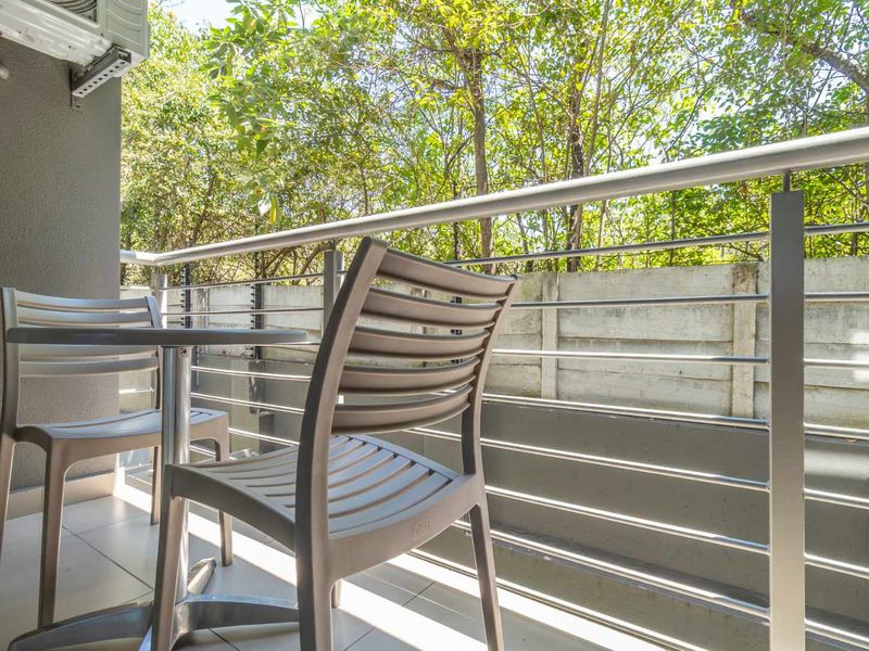 A private balcony featuring tiled flooring, a modern metal railing, and an external air conditioning unit, overlooking a lush green garden area.