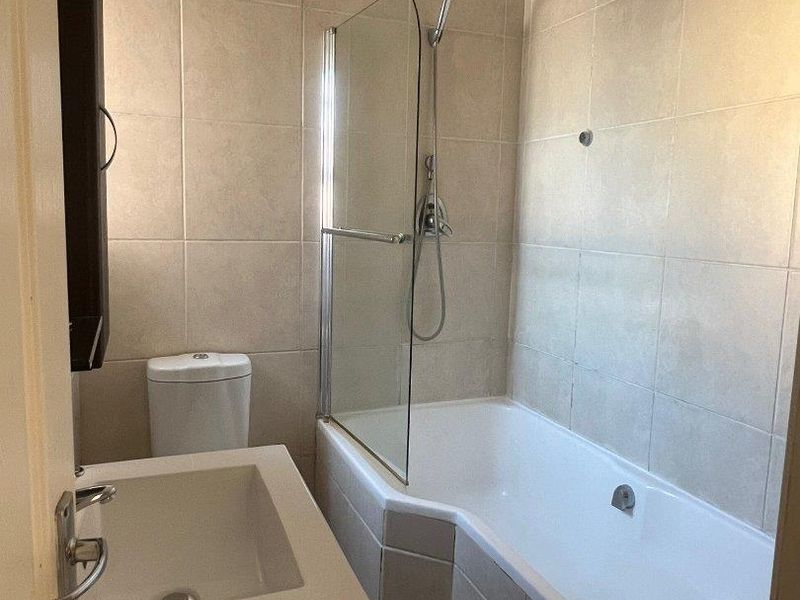 A clean, functional bathroom featuring a built-in vanity, toilet, and a bathtub with an integrated glass shower screen and neutral wall tiling.