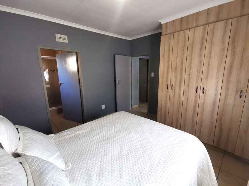 A well-maintained bedroom featuring light wood-finish built-in cupboards, neutral grey walls, tiled flooring, and direct access to an en-suite bathroom.