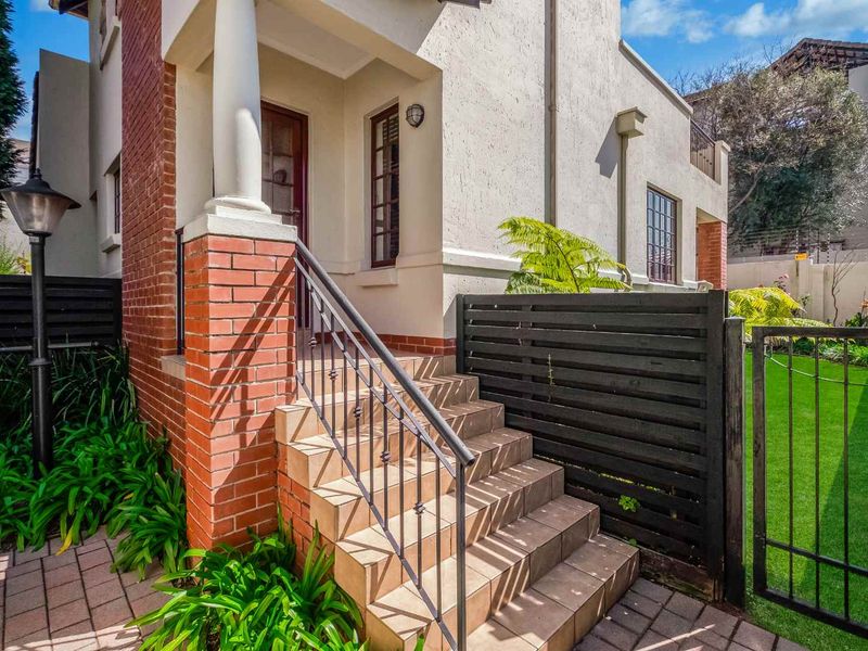 Front entrance of the townhouse featuring a brick facade, tiled staircase with wrought iron railing, and a well-maintained garden area with paved walkway.