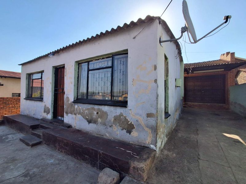 Front exterior view of a single-story house showing weathered plaster, peeling paint, security bars on windows, and a concrete entrance step.