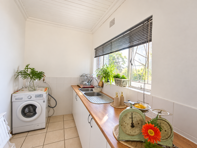 A functional laundry room featuring built-in cabinetry, a wooden countertop, stainless steel sink, and tiled splashback with natural light from the window.