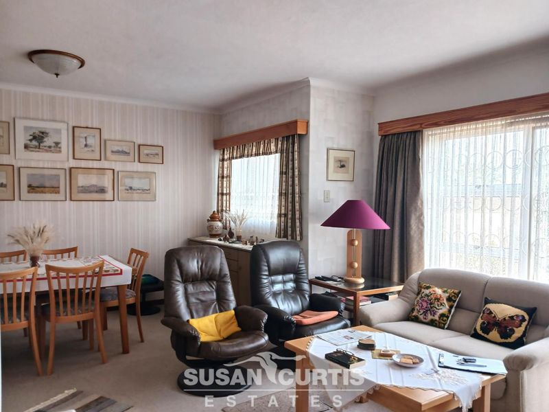 An open-plan living and dining area featuring neutral wallpaper, large windows with security bars, and classic ceiling lighting.