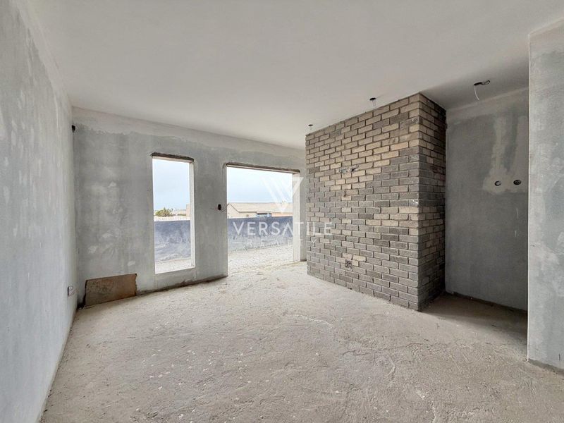 An unfinished living room space featuring an exposed brick wall, concrete flooring, and plastered walls, awaiting final finishes and electrical installation.