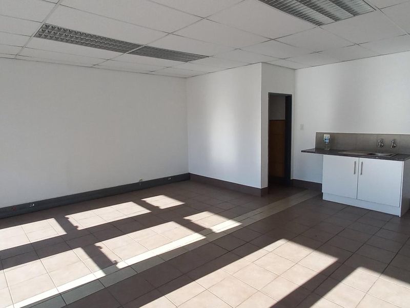 An open-plan industrial office space featuring tiled flooring, a suspended ceiling with recessed lighting, and a compact kitchenette area.