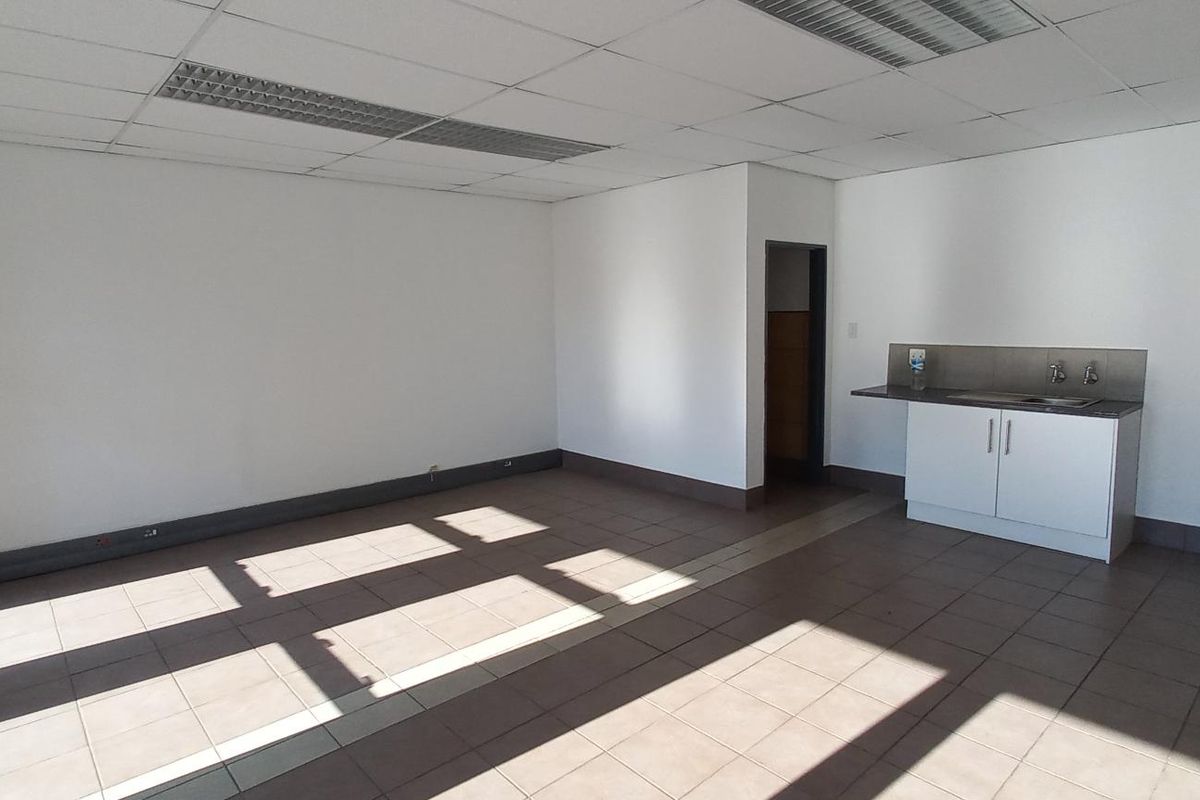 An open-plan industrial office space featuring tiled flooring, a suspended ceiling with recessed lighting, and a compact kitchenette area.