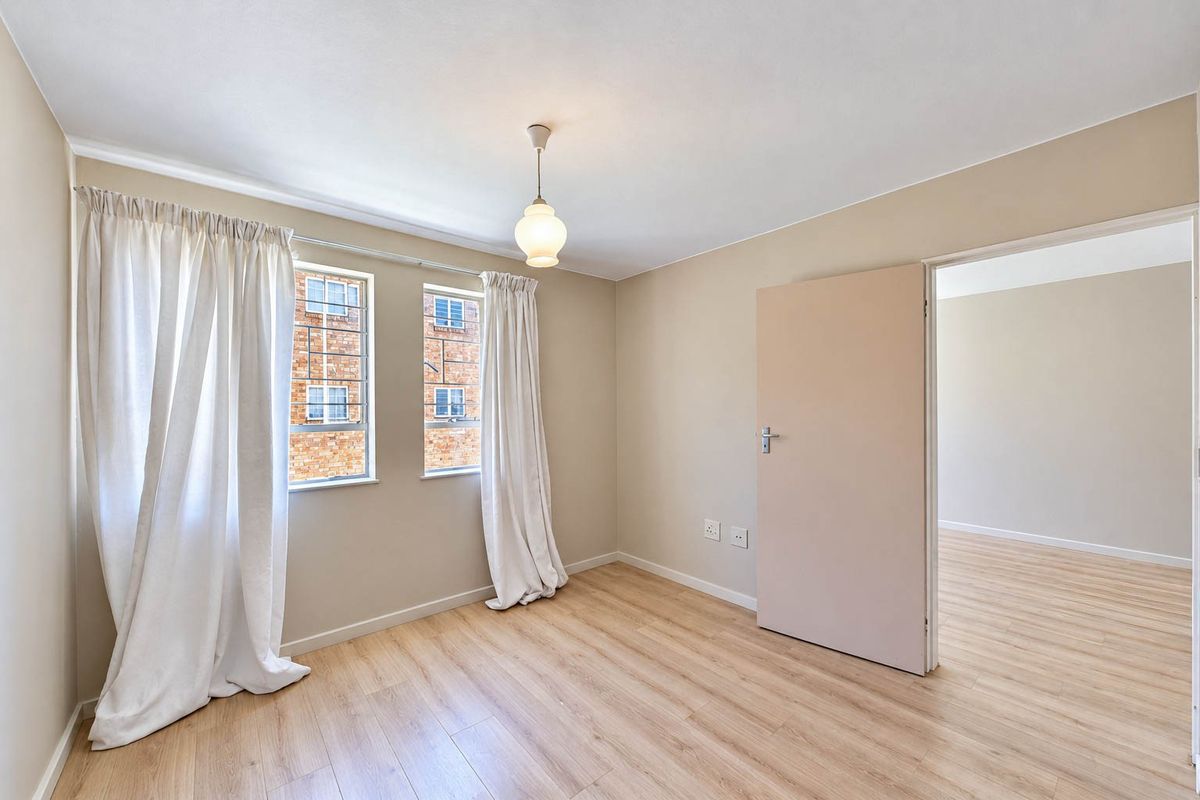 A bright, empty bedroom featuring light-toned laminate flooring, neutral walls, and security bars on the windows, offering a clean and versatile living space.