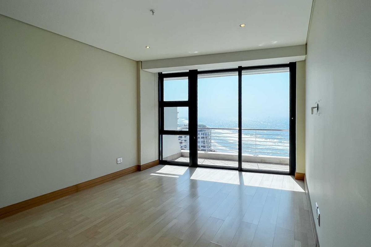 A bright, empty bedroom featuring light wood flooring, recessed ceiling lighting, and large sliding glass doors opening onto a balcony with ocean views.