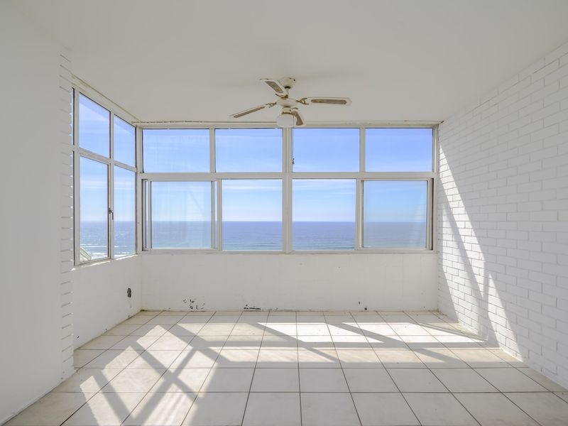 A bright, empty living area featuring white painted brick walls, tiled flooring, a ceiling fan, and expansive windows offering panoramic ocean views.