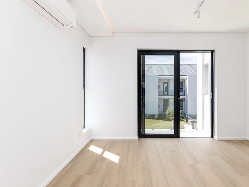 A bright, modern, and empty bedroom featuring light wood-look laminate flooring, a wall-mounted air conditioning unit, and sliding glass doors leading to an outdoor area.