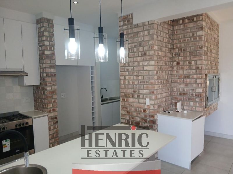 Modern open-plan kitchen featuring exposed brick accents, a central island, built-in braai, and contemporary cabinetry with integrated appliances.