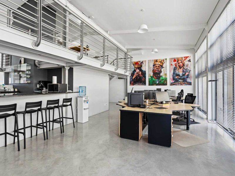 Modern open-plan office space featuring a mezzanine level, polished concrete floors, large windows with blinds, and a built-in kitchenette area.