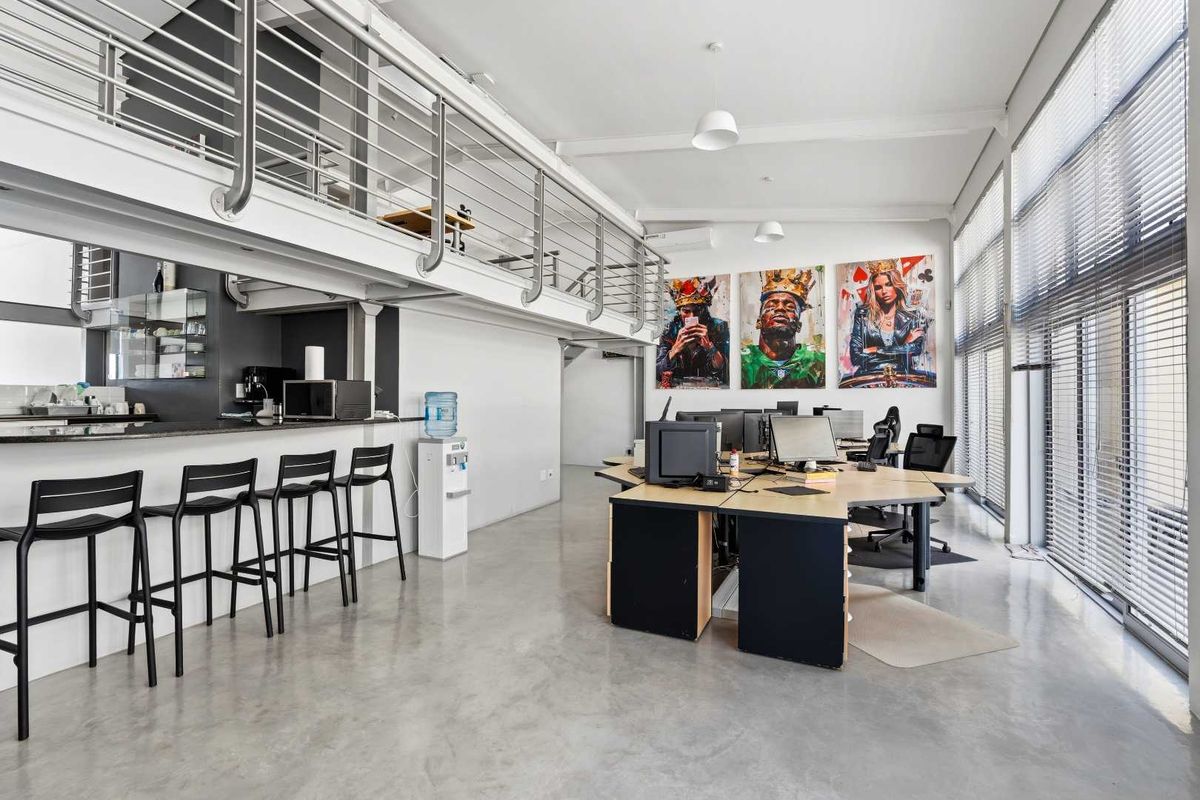 Modern open-plan office space featuring a mezzanine level, polished concrete floors, large windows with blinds, and a built-in kitchenette area.