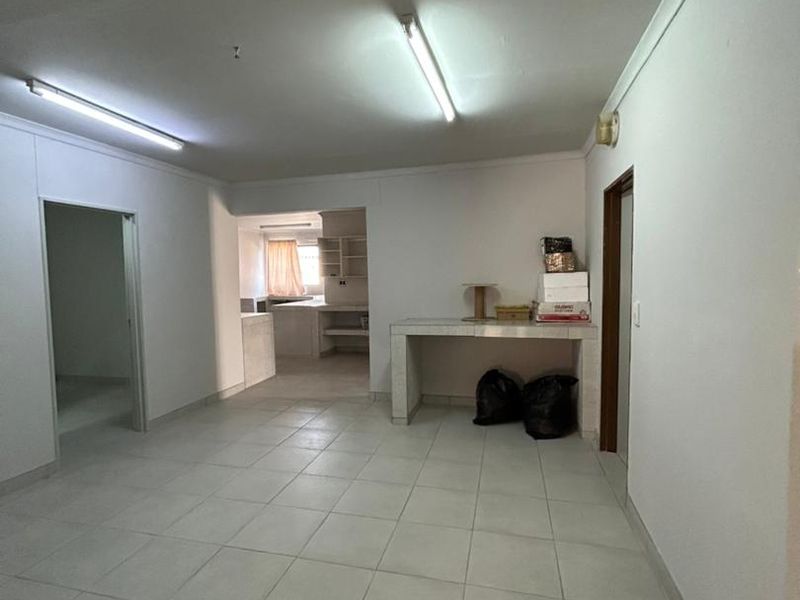 An open-plan living area featuring tiled flooring, fluorescent lighting, and a built-in stone counter, leading through to a secondary kitchen or utility space.
