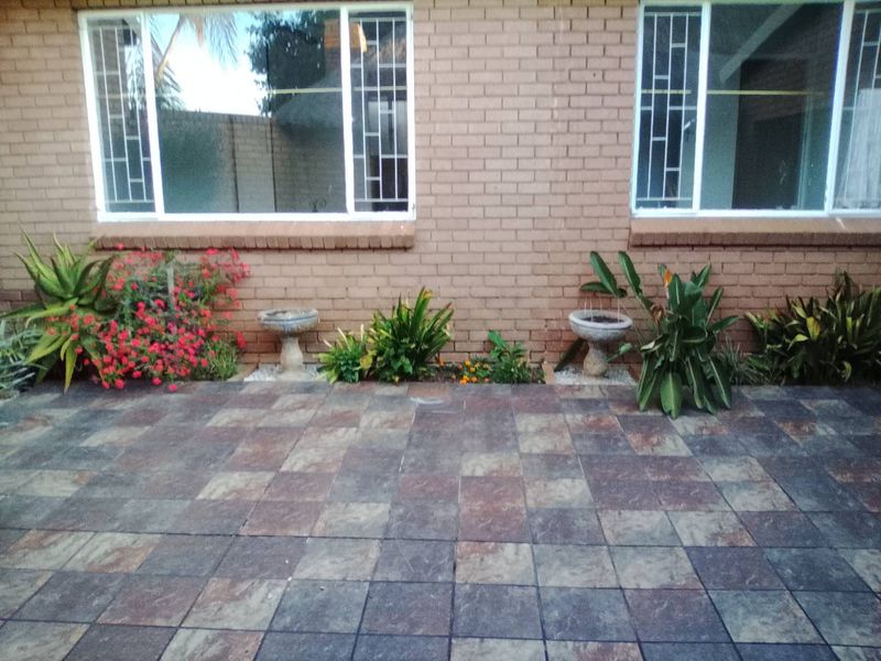 A rear exterior view featuring a paved patio area, brick wall facade, and windows fitted with security bars.