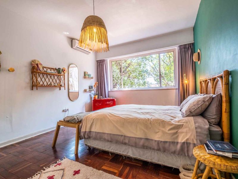 A well-lit bedroom featuring warm parquet flooring, a split-system air conditioning unit, and a large window providing natural light and garden views.