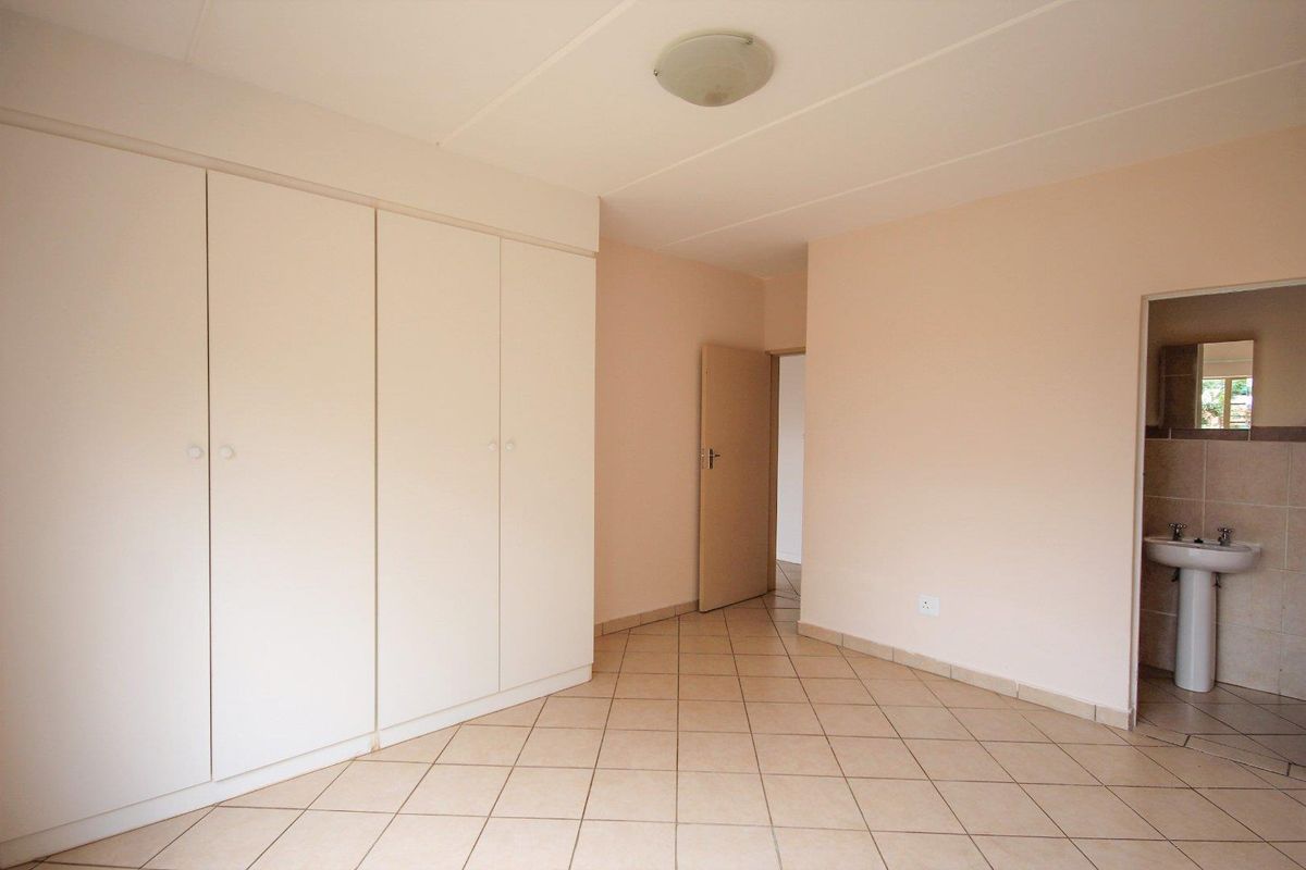 A spacious, empty bedroom featuring neutral tiled flooring, built-in cupboards, and an en-suite bathroom area with a pedestal basin.