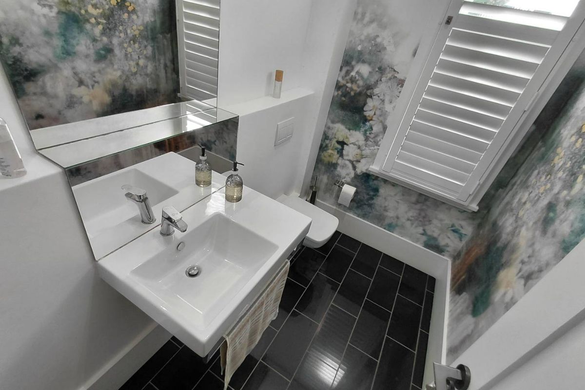 Modern bathroom featuring a wall-mounted basin, wall-hung toilet, sleek black floor tiles, and decorative wall finishes with plantation shutters.