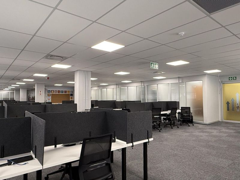 A spacious, modern open-plan office area featuring suspended ceilings, recessed LED lighting, carpeted floors, and glass-partitioned meeting rooms or private offices.