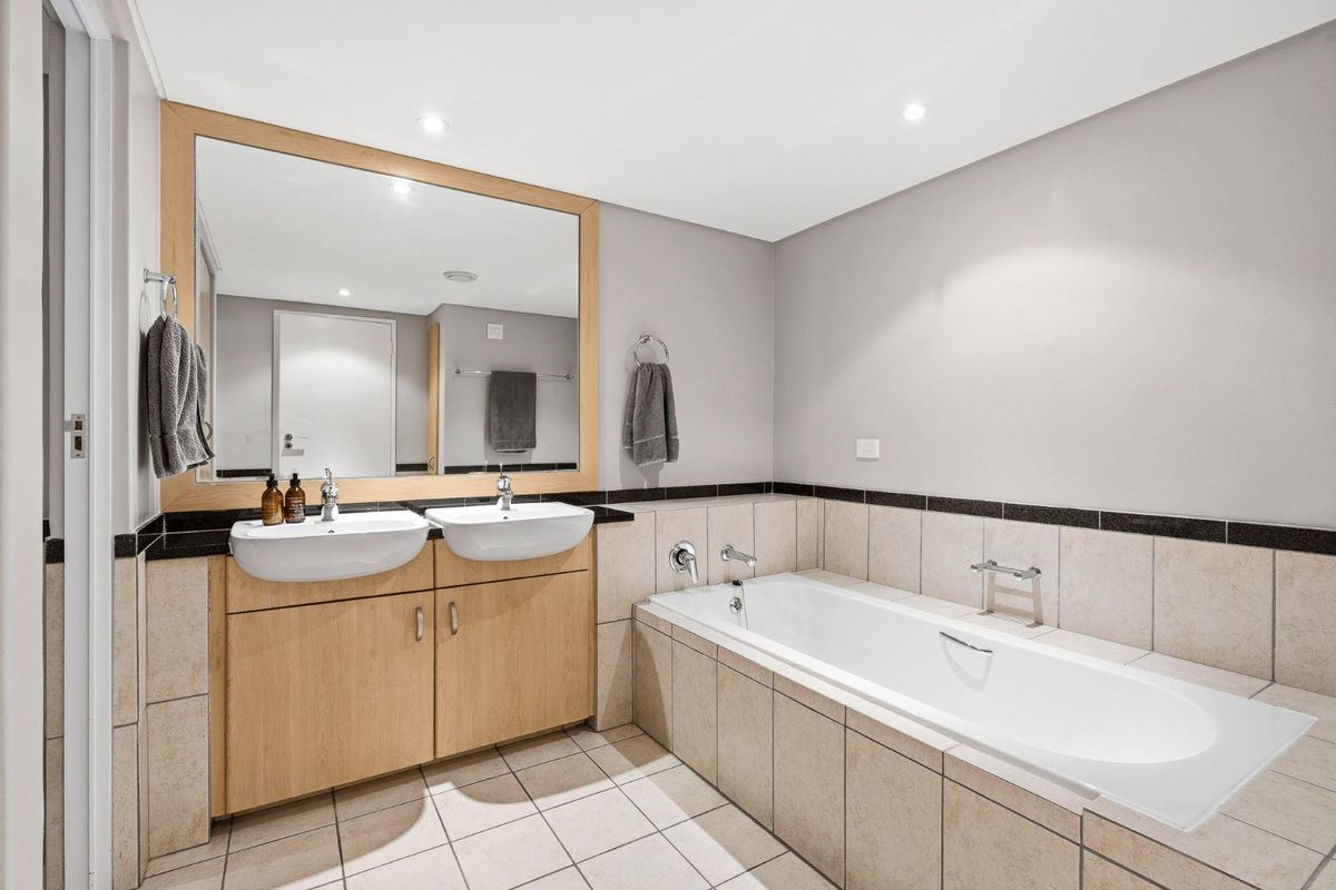 A well-maintained bathroom featuring a double vanity with wooden cabinetry, a large wall mirror, and a tiled built-in bathtub.