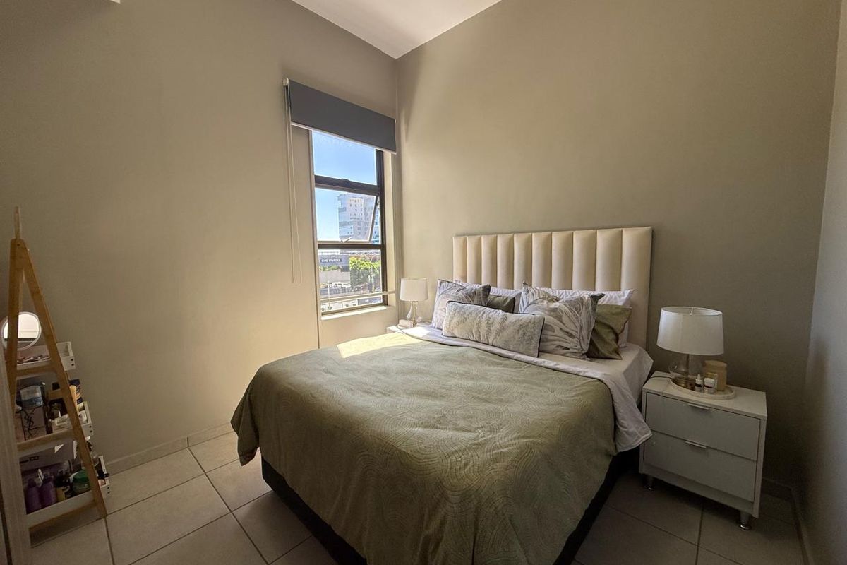 A well-lit bedroom featuring neutral wall tones, tiled flooring, and a large window with a roller blind, offering a comfortable living space.