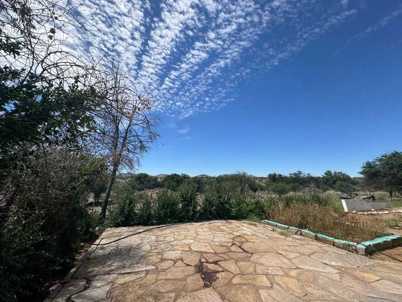 An outdoor stone-paved patio area set within a rural landscape, featuring a low masonry border and expansive views of the surrounding farm terrain.