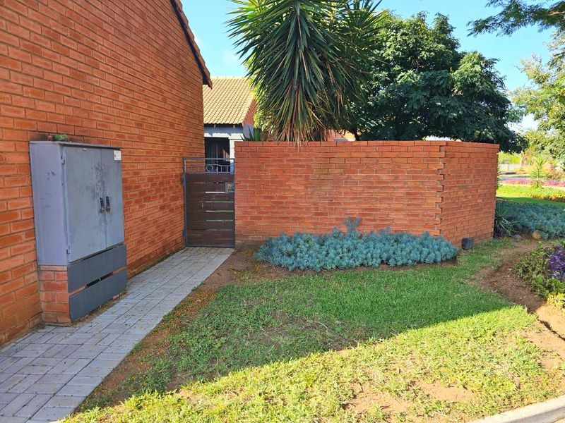 A view of the townhouse exterior featuring a brick boundary wall, a paved walkway leading to a pedestrian gate, and established garden landscaping.