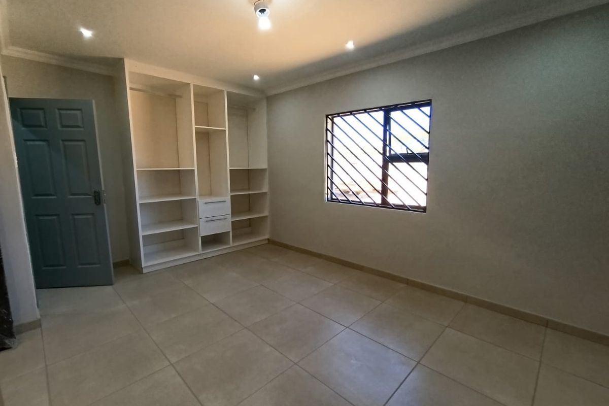 A bright, empty bedroom featuring neutral floor tiles, built-in white cabinetry, and a window secured with metal burglar bars.