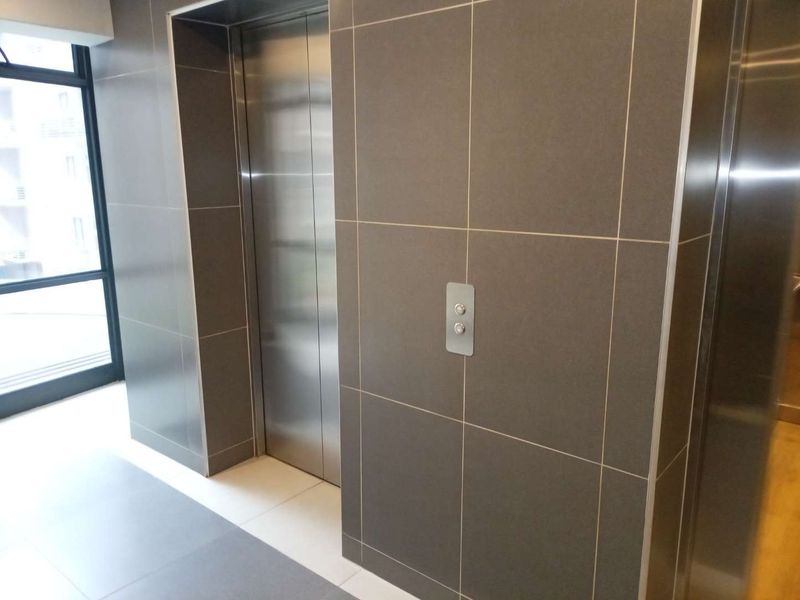 A modern building hallway featuring a stainless steel elevator, sleek dark grey tiled wall cladding, and large format floor tiles.