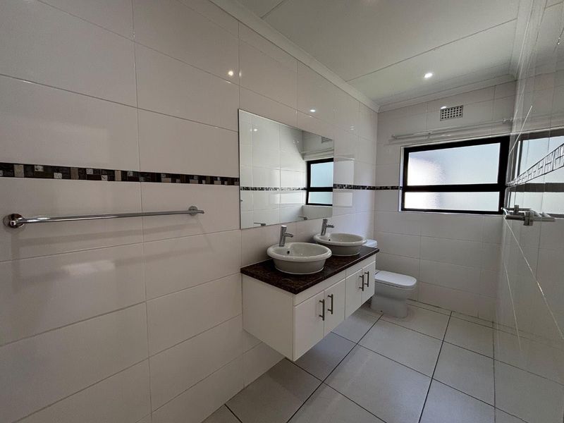 A clean, modern bathroom featuring a double vanity with vessel sinks, granite countertop, wall-mounted cabinetry, and neutral tiled finishes throughout.