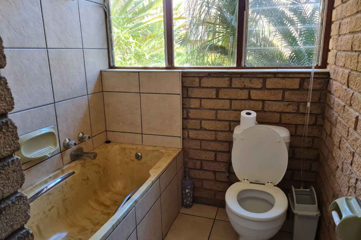 A dated bathroom featuring a built-in bathtub with tiled surround, a standard toilet, and exposed brick wall finishes.
