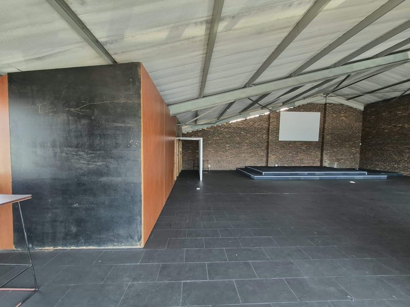 Spacious open-plan office area featuring exposed brick walls, steel roof trusses, tiled flooring, and a built-in stage platform suitable for presentations or events.