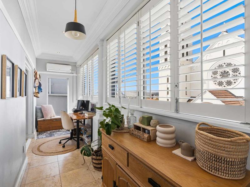 A bright, enclosed balcony or sunroom featuring tiled flooring, plantation shutters, and a split-system air conditioner, offering a pleasant space for a home office.