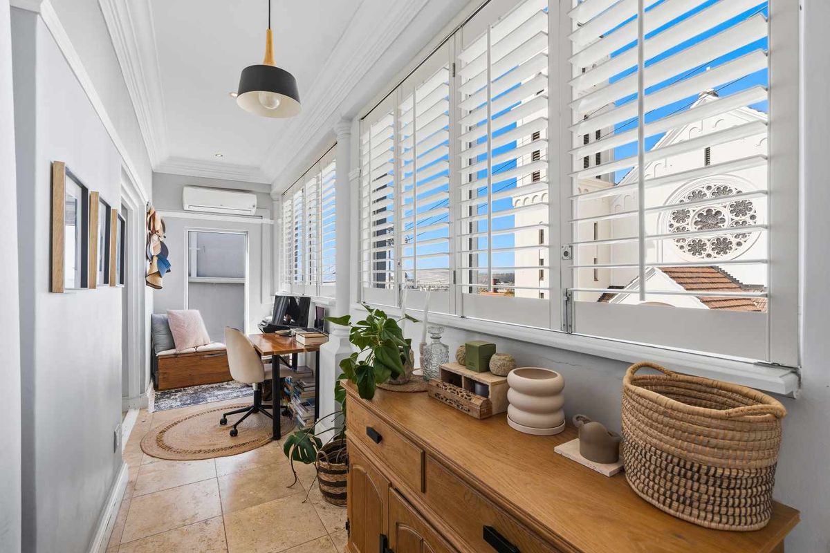 A bright, enclosed balcony or sunroom featuring tiled flooring, plantation shutters, and a split-system air conditioner, offering a pleasant space for a home office.