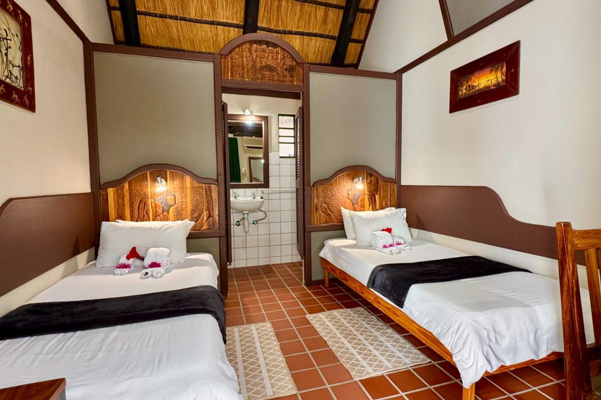 A twin bedroom featuring a thatched roof, tiled flooring, and an integrated en-suite bathroom with a wall-mounted basin.