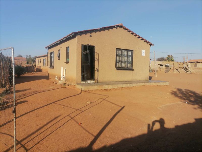 A detached single-story house featuring a tiled roof, security bars on windows, and a concrete porch, situated on an unpaved plot.
