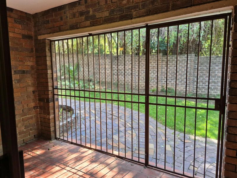 A covered patio area featuring exposed brick walls and tiled flooring, secured with a metal gate and overlooking a private garden space.