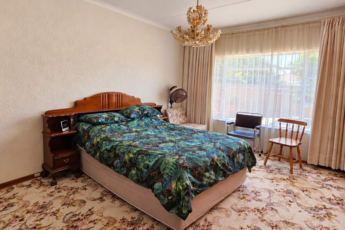A spacious bedroom featuring built-in wooden cupboards, large windows with security bars, and classic floral carpeting.