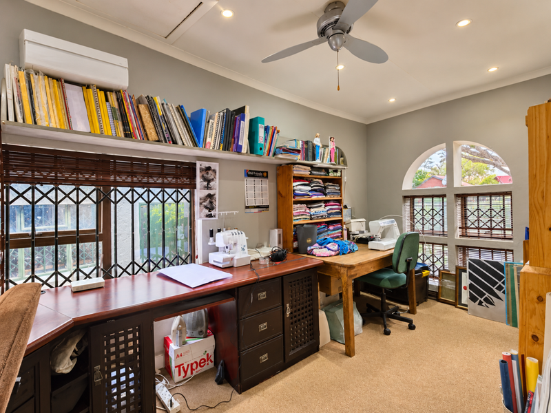 A functional home office featuring built-in shelving, a ceiling fan, split-system air conditioning, and arched windows with security gates.