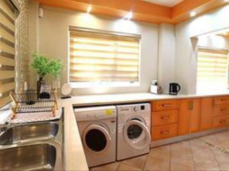A functional laundry room featuring built-in wooden cabinetry, laminate countertops, a double stainless steel sink, and tiled flooring with recessed lighting.