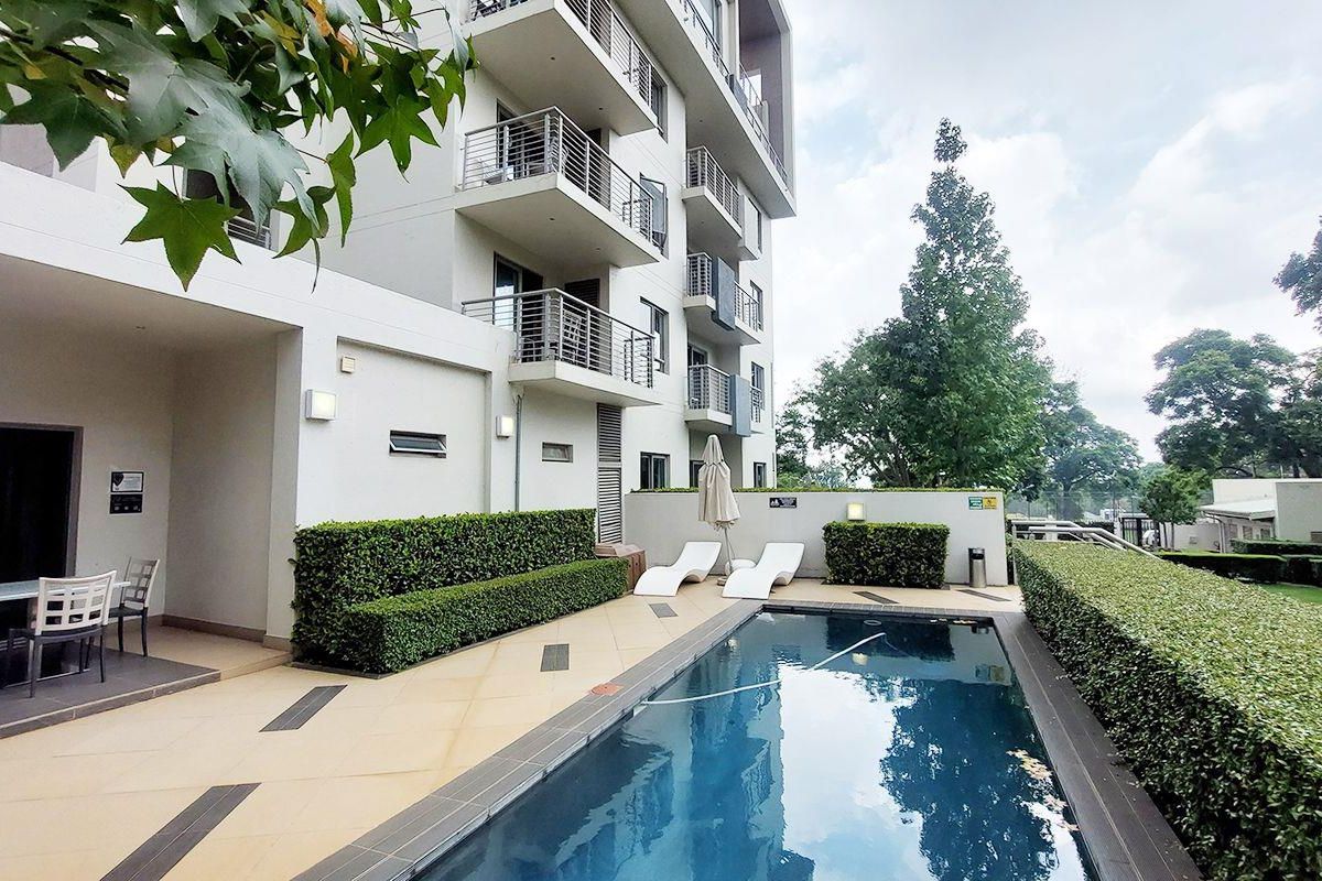 A well-maintained communal swimming pool area featuring a tiled deck, manicured hedges, and direct access to the apartment building's exterior balconies.