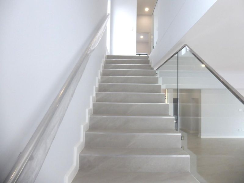 A modern, minimalist staircase featuring tiled steps, a sleek glass balustrade, and a wall-mounted handrail in a bright, open hallway.