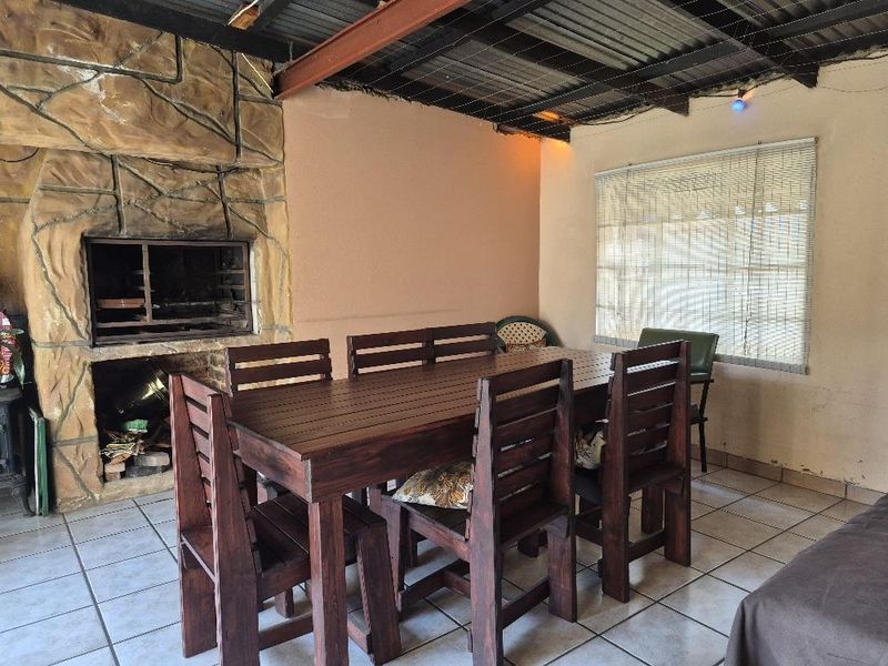 Covered patio area featuring a built-in stone-clad braai, exposed beam ceiling, and tiled flooring, suitable for outdoor dining and entertainment.