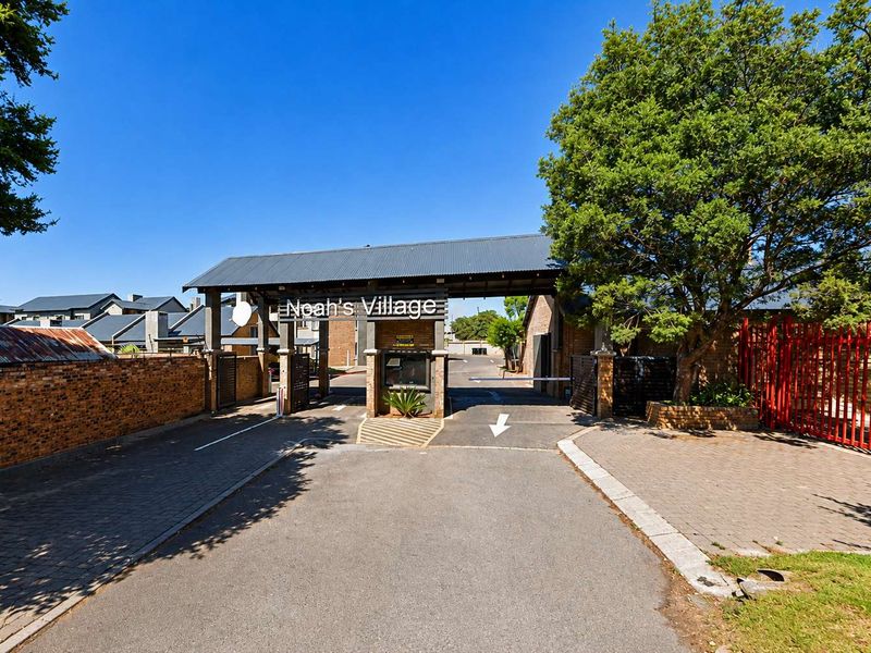 The entrance to Noah's Village featuring a secure gatehouse, boom gate access, and paved driveway surrounded by established trees and brick boundary walls.