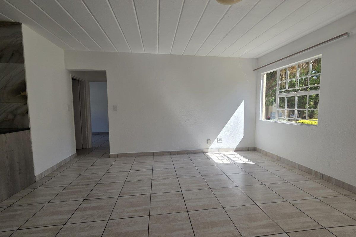 A bright, empty living area featuring neutral tiled flooring, a panelled ceiling, and large windows allowing for ample natural light.