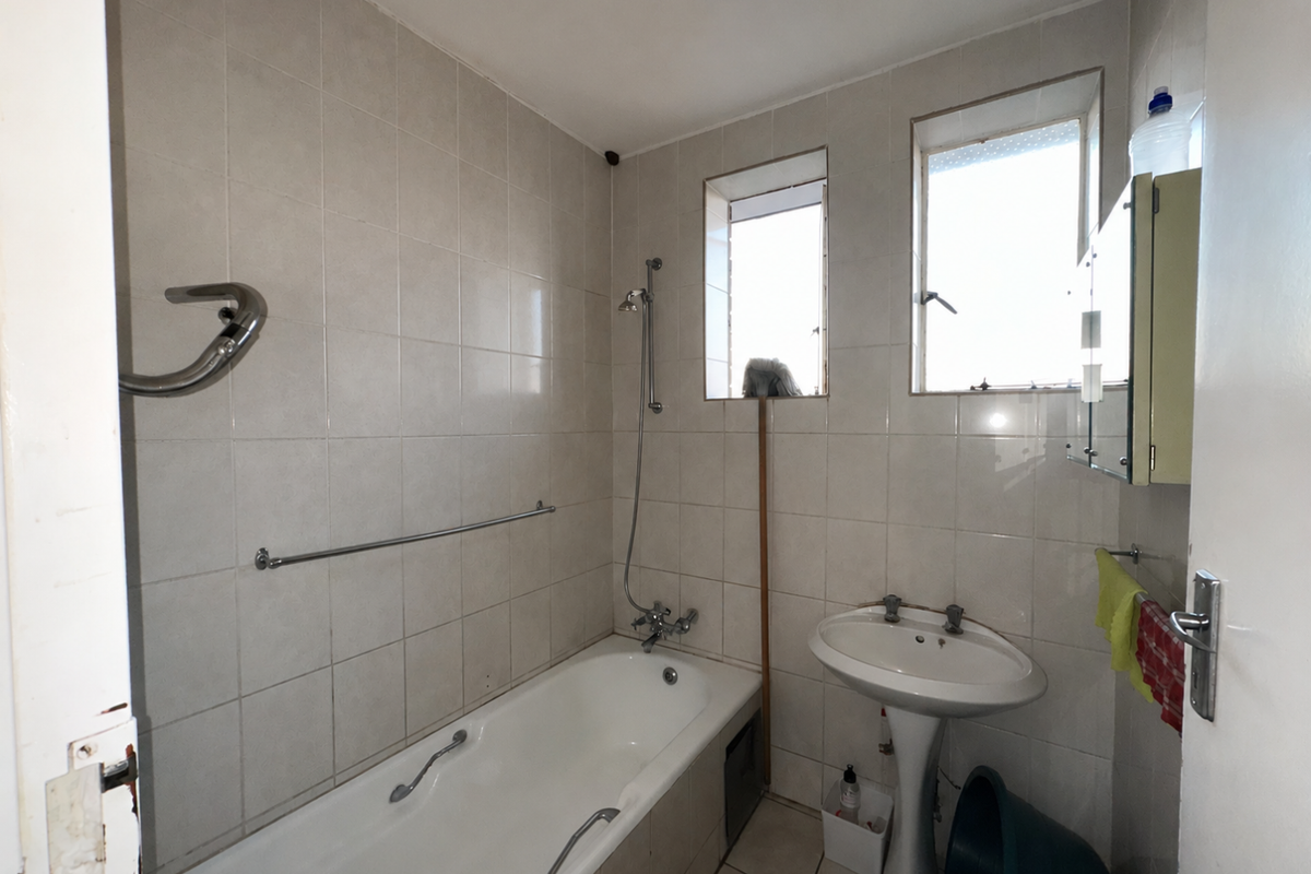 A dated bathroom featuring a standard bathtub with shower mixer, pedestal basin, and tiled walls, requiring cosmetic updates to modernize the space.
